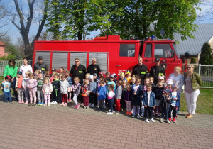 Dzieci stoją pośród strażaków, w tle wóz strażacki.